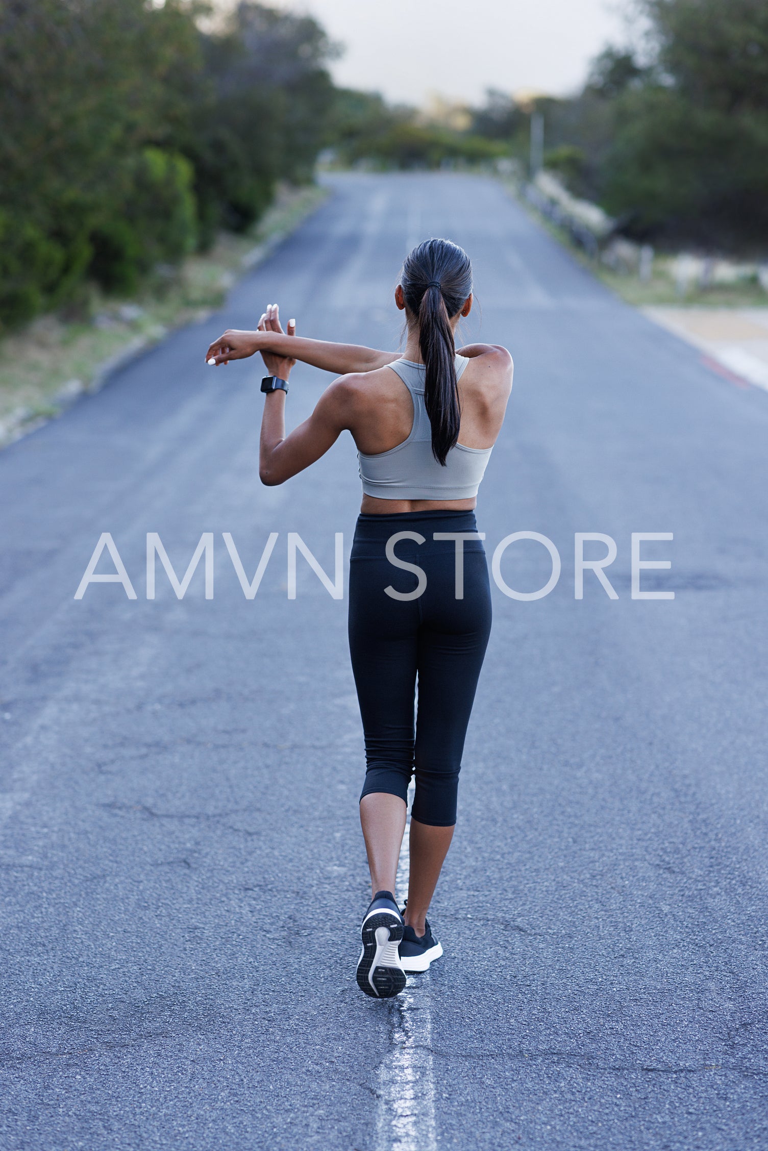 Full length of slim athlete walking on an abandoned road and warming up. Back view of female in fitness attire stretching her hands while walking.