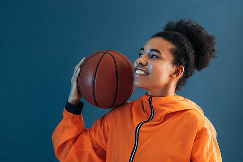 Smiling woman in orange sports clothes holding a basketball on her shoulder. Young female basketball player posing over blue backdrop.