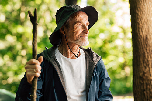 Portrait of a senior man with a wooden stick looking away while standing in the forest. Mature male wearing a hat and hiking clothes and looking away.