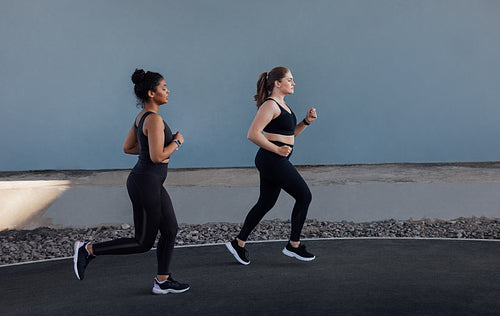 Two women with different body types running outdoors. Side view of two young females jogging at a grey wall.