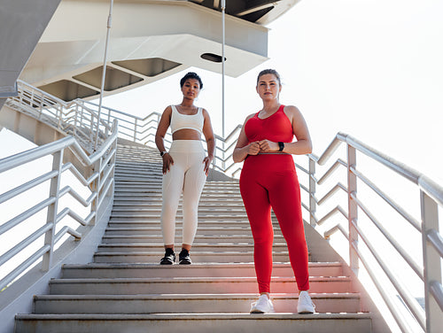 Two females in fitness attire with different colors looking at camera while standing on a staircase. Plus-sized woman with her fitness friend posing together.