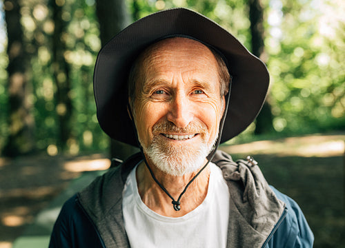 Close-up portrait of happy senior tourist looking at camera. Smiling male with a beard and hat looking at camera while standing in a forest during hiking.