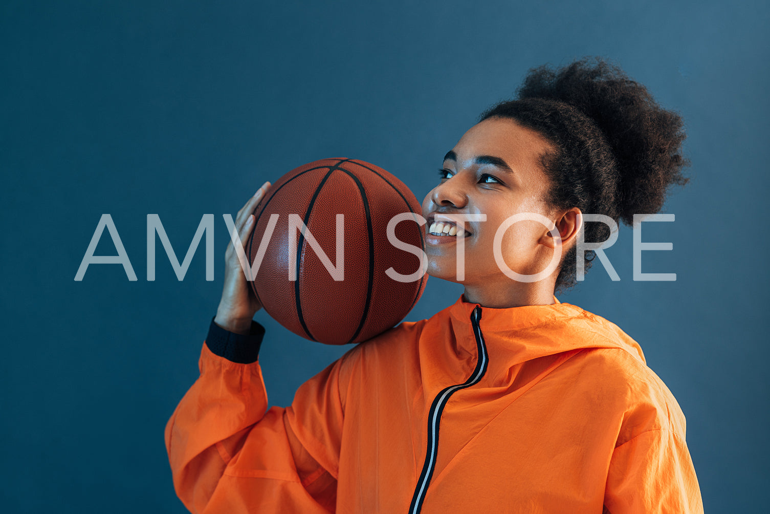 Smiling woman in orange sports clothes holding a basketball on her shoulder. Young female basketball player posing over blue backdrop.