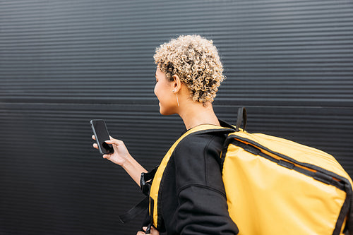 Side view of confident delivery woman with bag standing on a sidewalk holding a smartphone