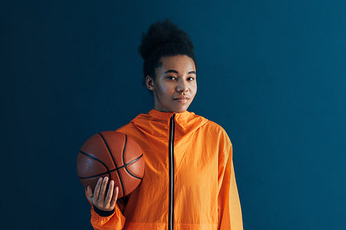 Portrait of young female basketball player against blue backdrop. Woman in orange sportswear holding a basketball looking at camera.