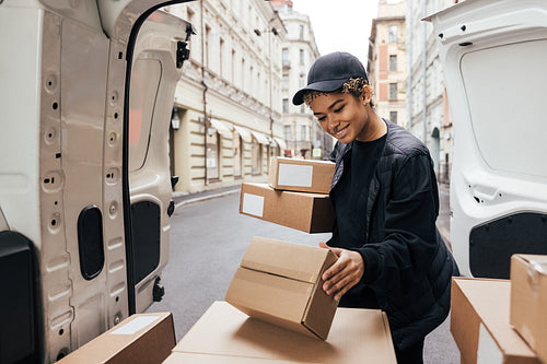 Smiling woman courier in uniform standing at car trunk holding b