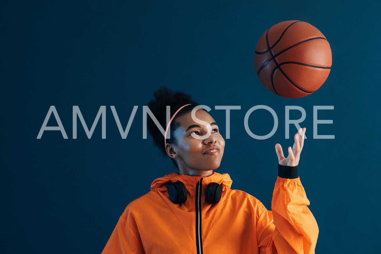 Cheerful female in orange fitness attire throw a basketball over blue background. Smiling sportswoman looking at flying basketball.