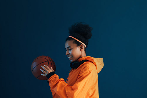 Smiling sportswoman in orange clothes holding a basketball. Side view of a young female with headphones holding basketball over a blue backdrop.