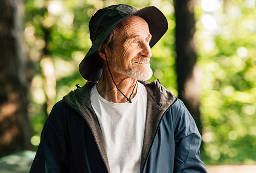 Portrait of a smiling senior tourist wearing a hat. Positive male tourist looking away while standing in a sunny forest.