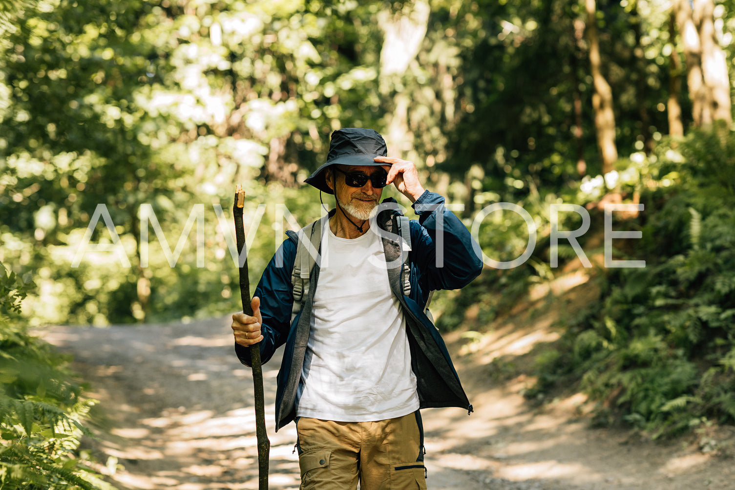 Senior male with a wooden stick wearing hat and sunglasses while trekking in forest