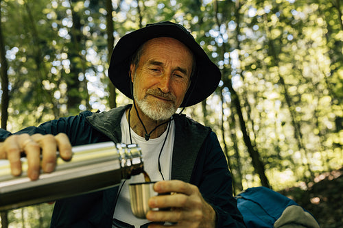 Senior male in hat pouring coffee from a thermos while taking a break in a forest during walk