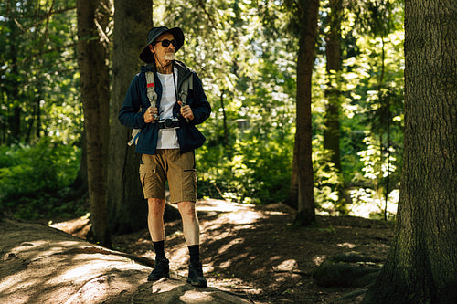 Confident senior tourist standing with backpack in forest and looking away