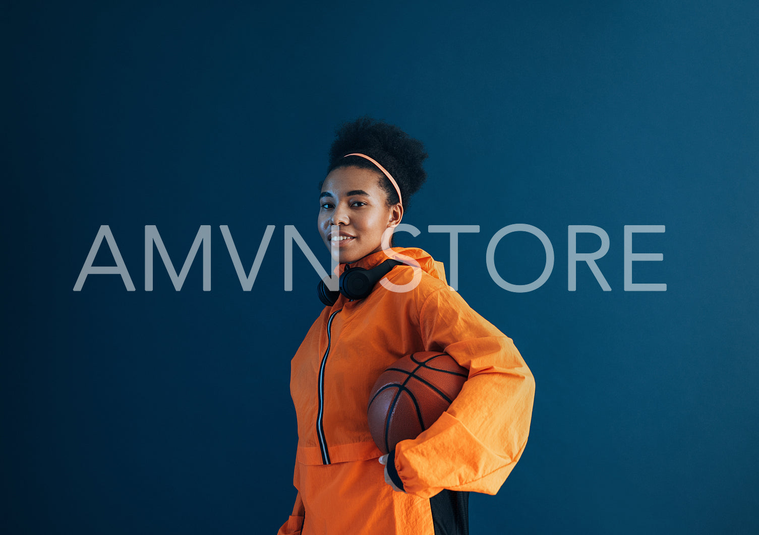 Young sportswoman standing with basketball in studio and looking at camera. Female in orange sportswear over blue background.