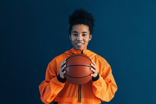 Young woman in orange fitness attire posing over a blue background. Female with basketball looking at camera in studio.