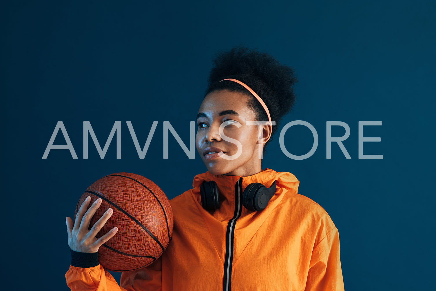 Young female with basketball and wireless headphones posing in studio. Confident sportswoman in orange fitness attire over blue background looking away.