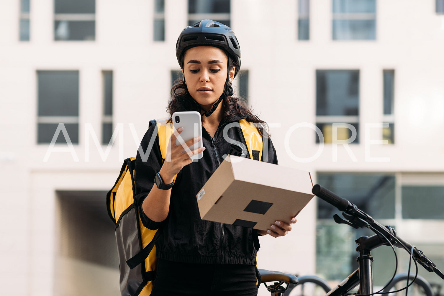 Delivery woman in cycling helmet holding a cardboard box checking information on mobile phone