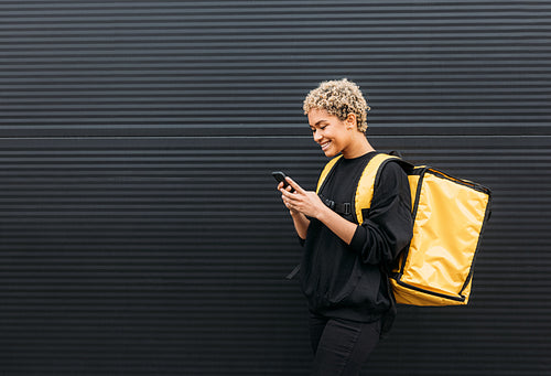 Smiling courier girl with food delivery backpack holding a smartphone while standing at black wall outdoors