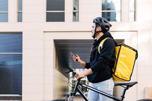 Side view of a young male working for a delivery company and looking for customer address. Courier standing at building with a thermal backpack.