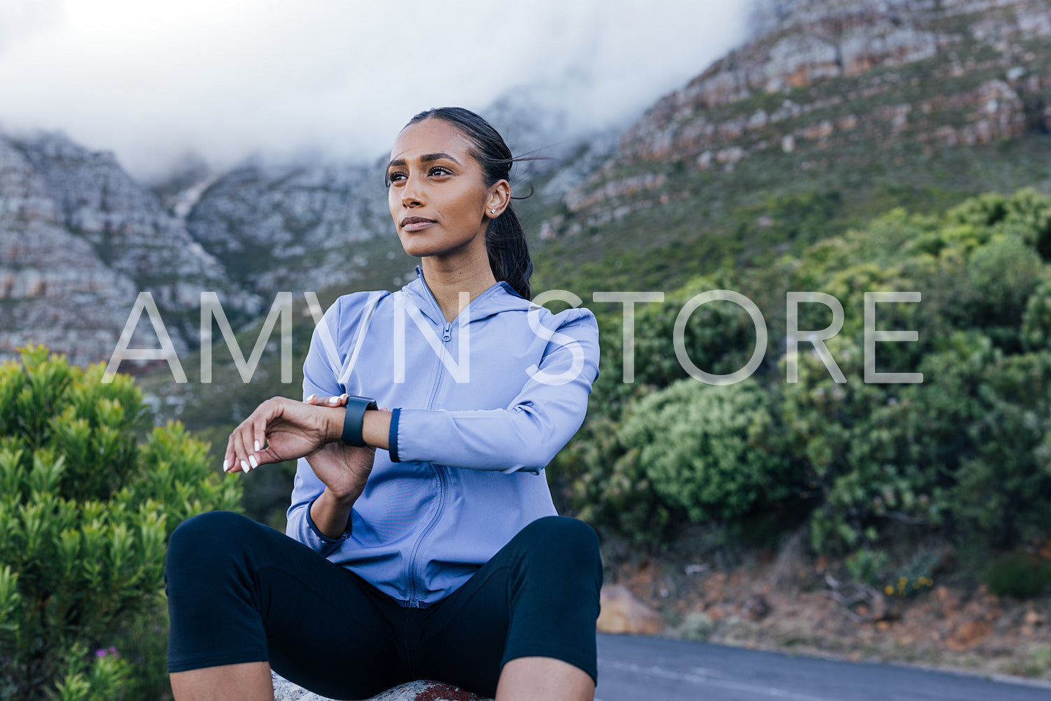 Slim woman in fitness attire sitting on the rock. Female using smartwatches looking into the distance.