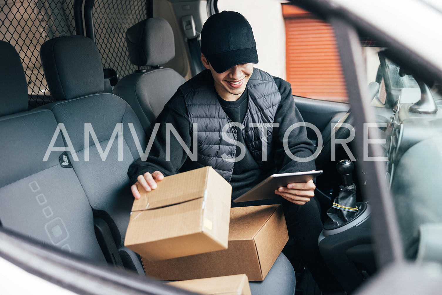 Smiling male courier in uniform and cap checking information on cardboard box while sitting in a car using a digital tablet