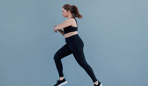 Side view of plus size female running against a grey wall wearing black fitness attire. Young woman with plus size body type jogging and looking at her smartwatch.