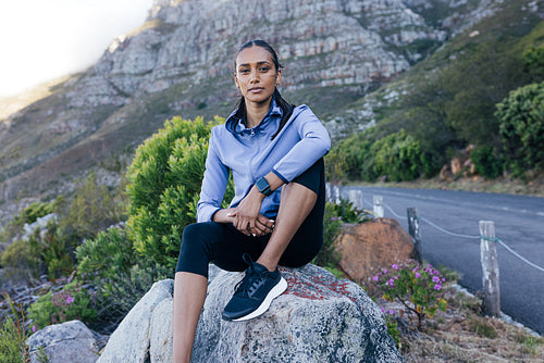 Young slim female sitting on rock relaxing after a trail run. Woman in sportswear looking at camera while sitting in natural park.