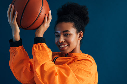 Close-up portrait of a young smiling basketball player looking at the camera over a blue backdrop. Female with curly hair with basketball wearing orange sportswear.