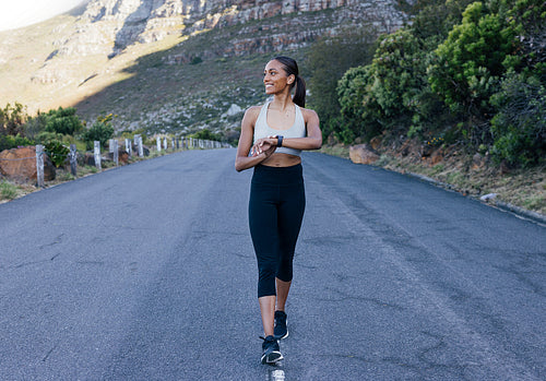 Slim smiling woman walking in the middle of the abandoned road checking her smartwatches. Full length of a young cheerful sports female looking away while walking on the road against a mountain.