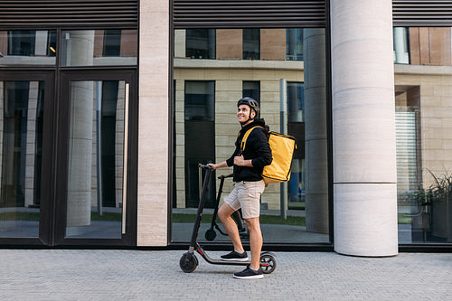 Smiling messneger man standing outdoors with electric push scooter. Delivery man with termal backpack in the city.