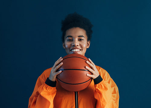 Portrait of a young female with curly hair holding a basketball and smiling against a blue backdrop