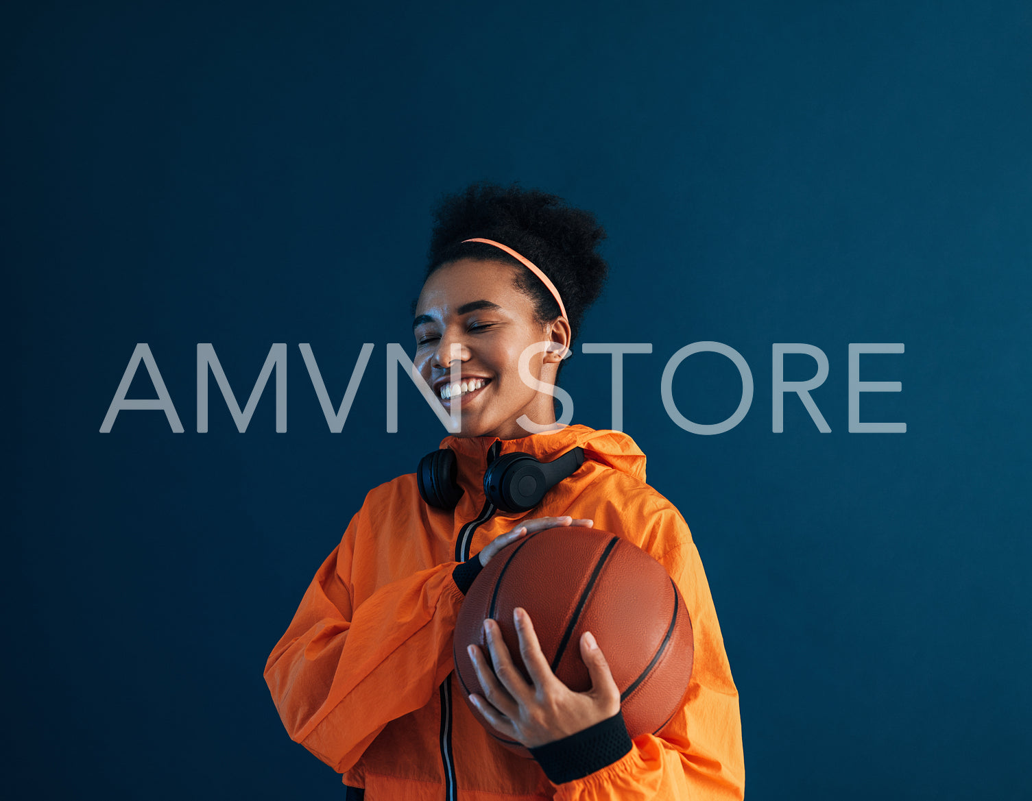 Happy sportswoman with closed eyes holding basketball while standing at blue backdrop in studio. Cheerful female posing with basketball in studio.