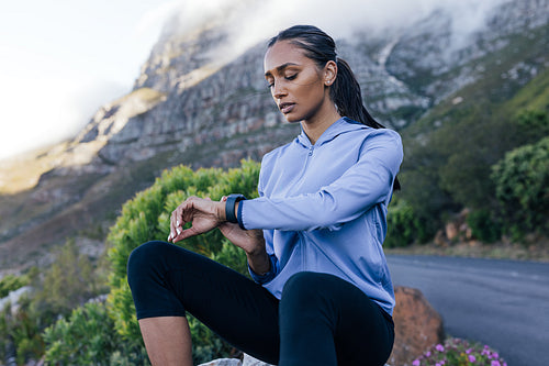 Young sportswoman checking pulse while sitting on a rock in a natural park. Female looking at smartwatch while sitting outdoors in a natural park.