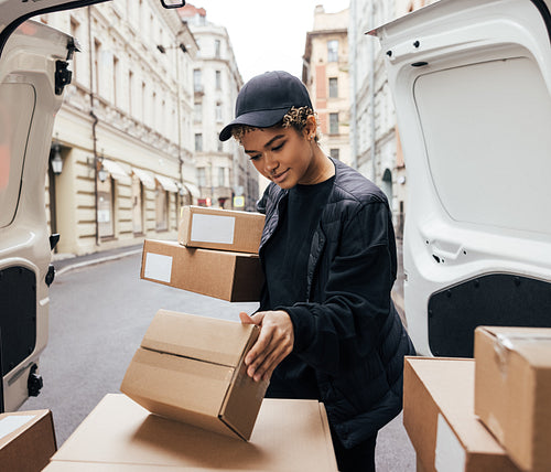 Young woman courier checking information on parcel while standing at van trunck