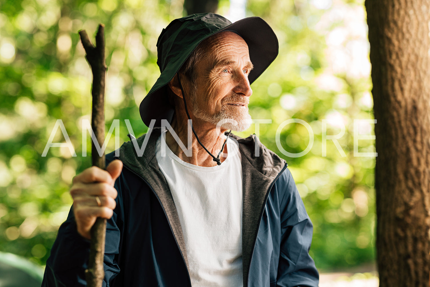 Portrait of a senior man with a wooden stick looking away while standing in the forest. Mature male wearing a hat and hiking clothes and looking away.