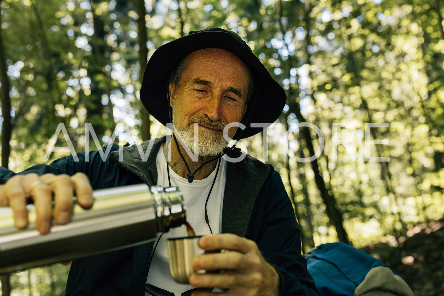 Senior male in hat pouring coffee from a thermos while taking a break in a forest during walk