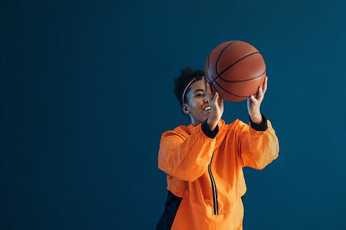 Smiling female basketball player throws basketball over blue backdrop in studio. Young woman in orange fitness wear practicing with basketball.