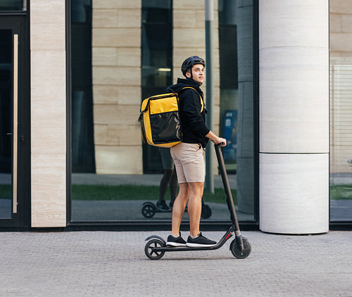 Side view of male courier riding on electric push scooter with food delivery backpack