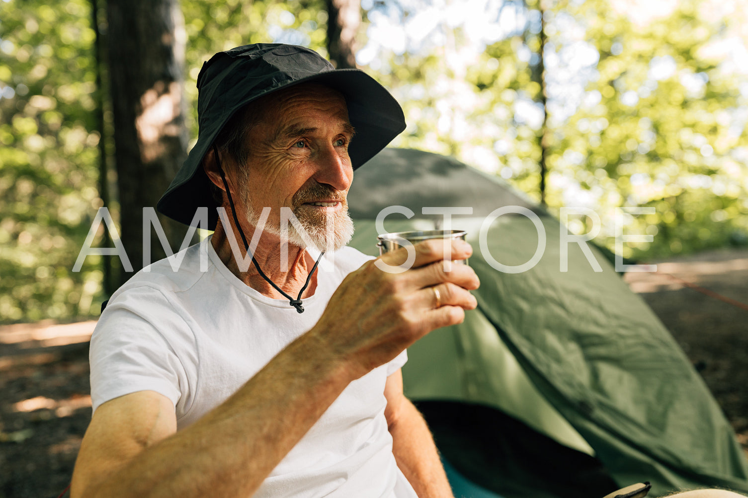 Senior male in hat holding metallic cup while sitting in the forest against tent