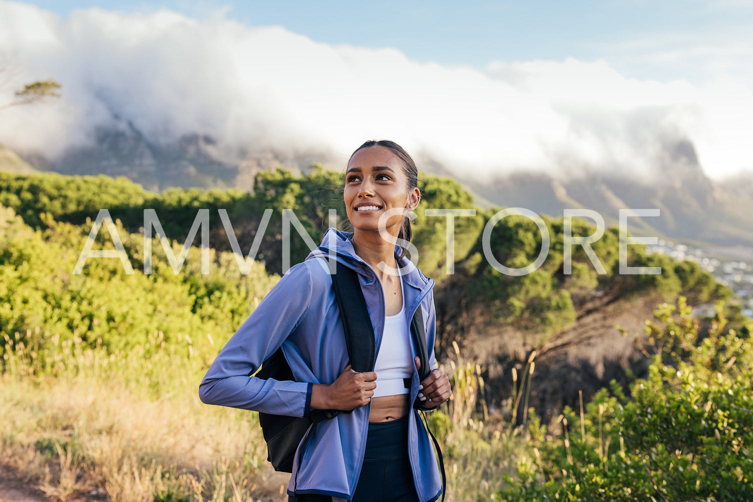 Smiling woman with backpack and sportswear enjoying hiking at sunset. Cheerful woman looking away while walking outdoors in a natural park.