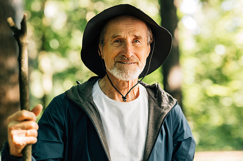 Portrait of a senior male wearing a hat and holding a stick. Mature man looking at the camera during his forest walk.