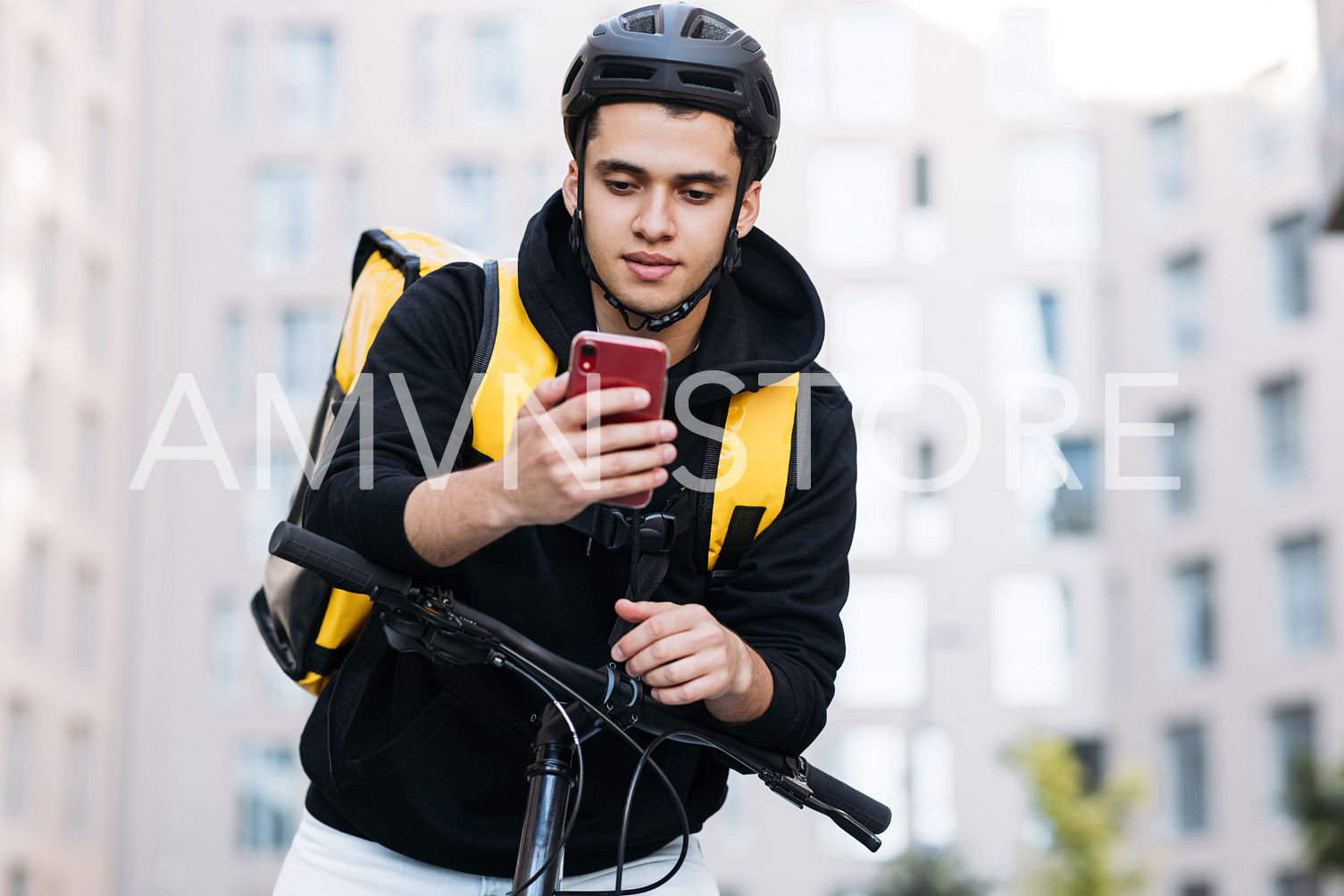 Male courier leaning on the handlebar of a bicycle wearing food delivery backpack and cycling helmet looking at mobile phone