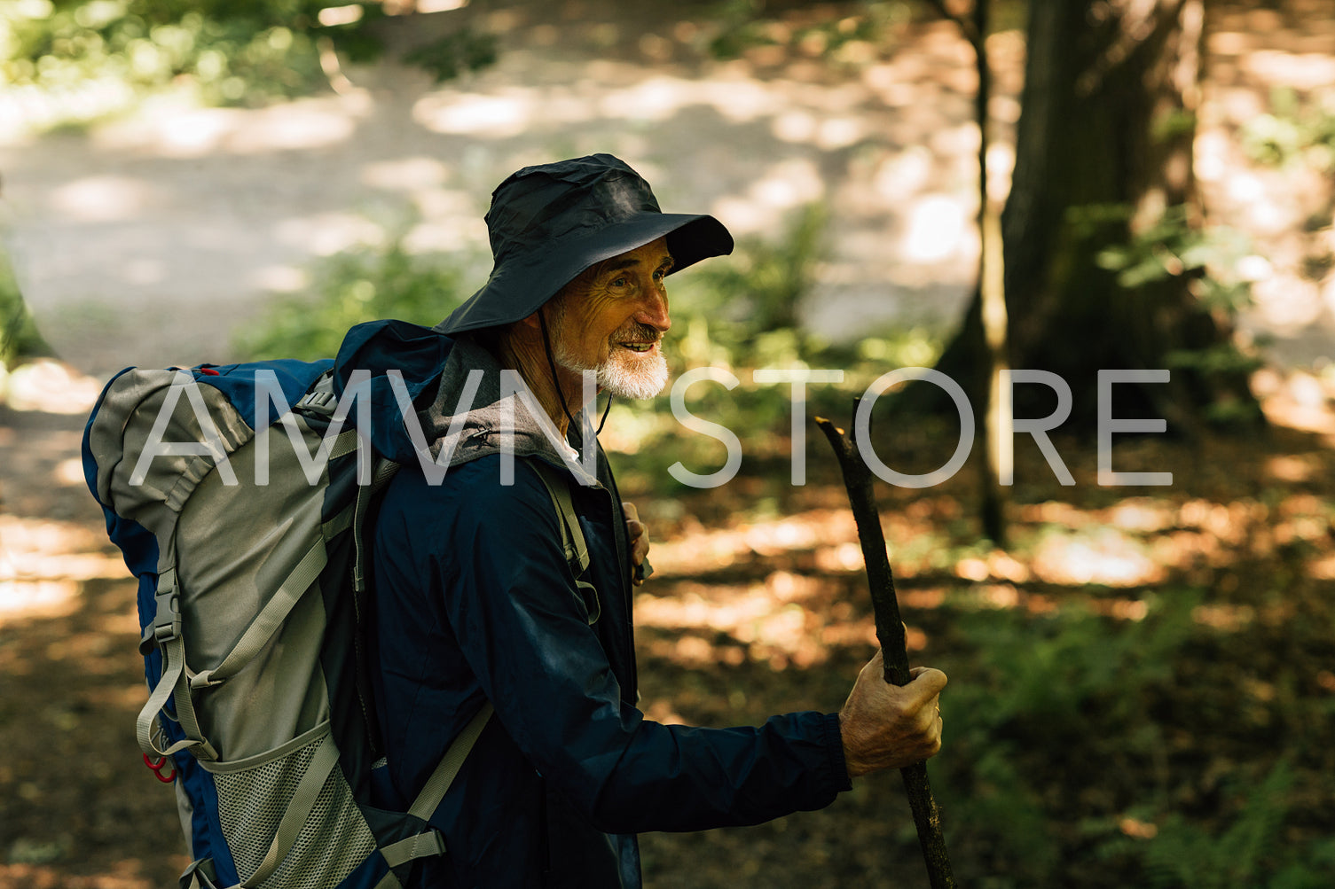 Cheerful senior male with a wooden stick and tourist backpack walking in a forest