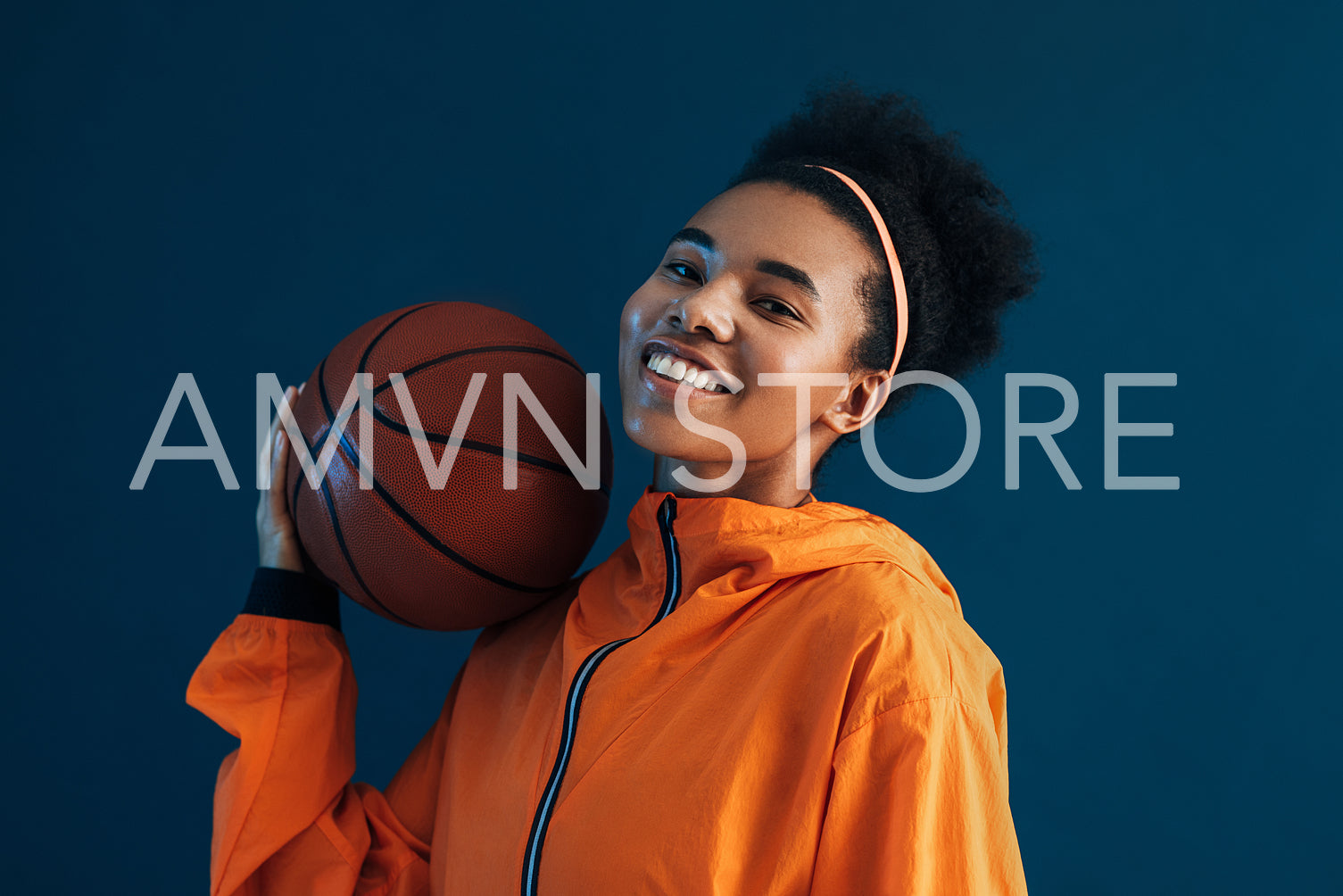 Cheerful basketball player posing with basketball in the studio over blue backdrop. Smiling female in orange sportswear.