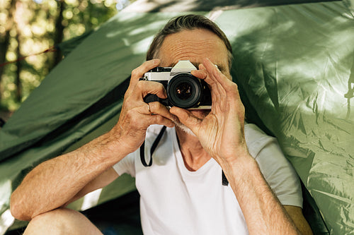 Senior male photographing while sitting in the forest during a hike. Mature man with a film camera sitting a tent outdoors.