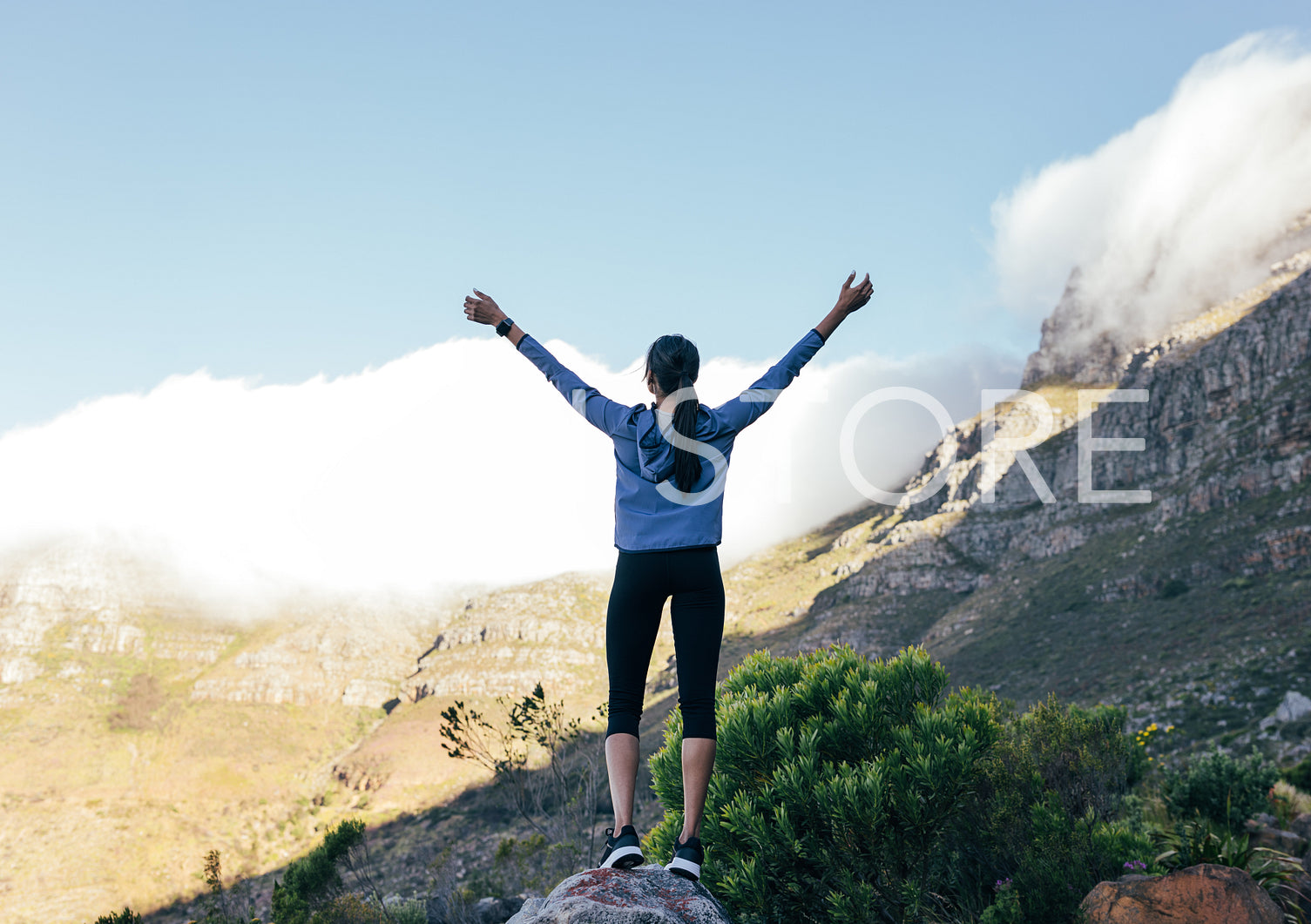 Back view of young slim female raising her hands in front of a mountain with clouds during a trail run. Full length of sportswoman standing on rock and looking at mountains.