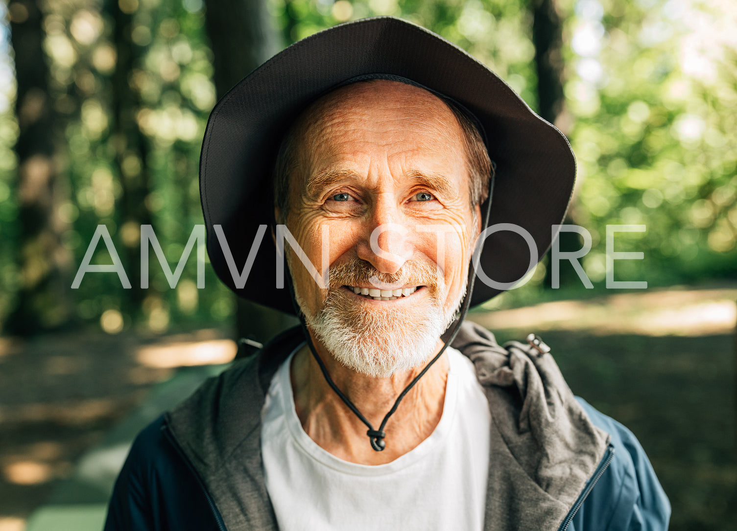 Close-up portrait of happy senior tourist looking at camera. Smiling male with a beard and hat looking at camera while standing in a forest during hiking.