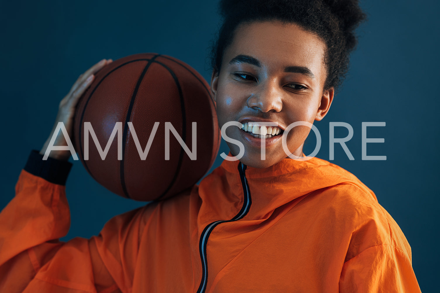 Close-up of young professional female basketball player over blue backdrop. Cheerful woman in orange sportswear with basketball on her shoulder.