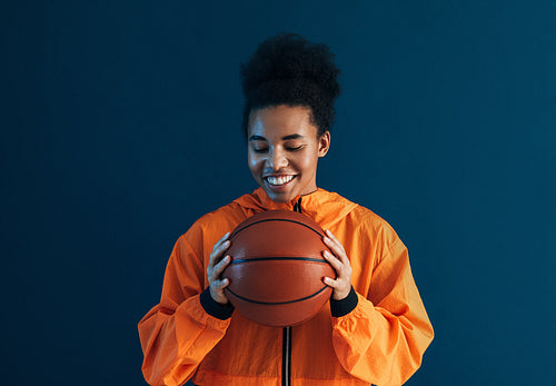 Positive female in orange sportswear holding a basketball. Young woman with closed eyes over blue backdrop holding a basketball.