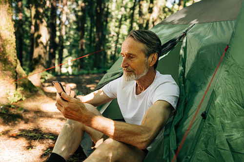 Mature male sitting in a tent and using a smartphone. Senior man with a smartphone in the forest.