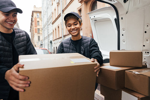 Two smiling coworkers unloading cardboard boxes from van for del